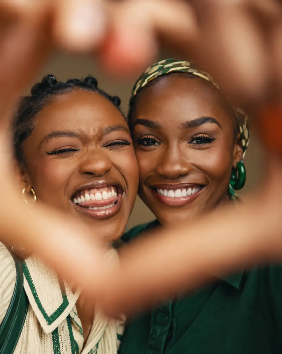 Two friends laughing cheek to cheek, making a heart shape with their hands towards the camera