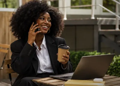 An employee smiling on a phone call at an outdoor café table with a coffee and laptop