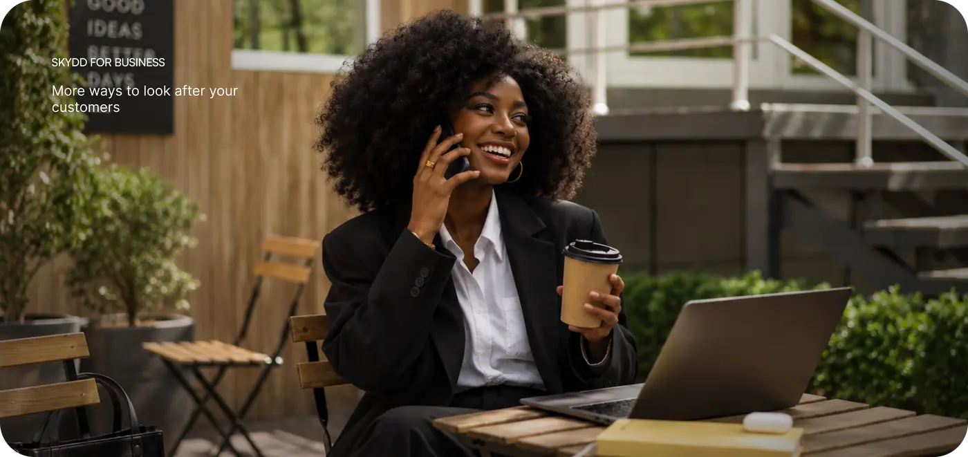 Skydd for Business — more ways to look after your customers. A smiling businesswoman on a phone call at an outdoor café table with a coffee and laptop.