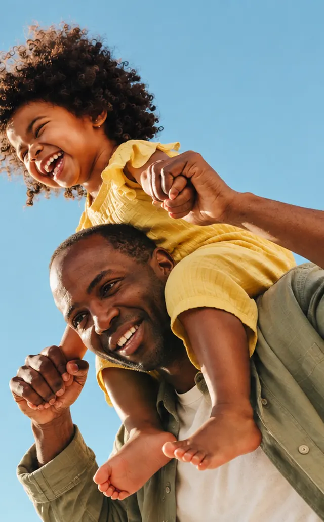 A laughing little girl riding on her smiling father's shoulders under a clear blue sky
